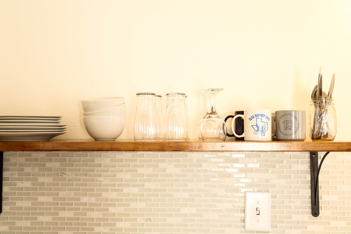 A wooden shelf displays a collection of plates, glasses, and mugs alongside a jar of utensils. The arrangement includes both clear and patterned glassware, providing a functional kitchen setup. A light-colored wall and a textured backsplash complete the setting.