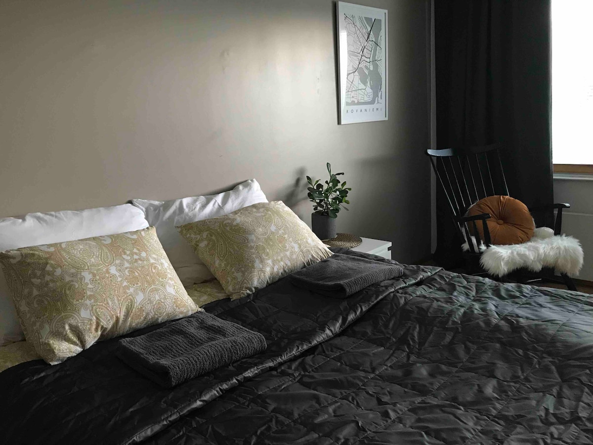 The sleeping area features a spacious bed with patterned pillows and folded towels positioned neatly on top. A cozy rocking chair is placed beside the window, accompanied by a small plant for added decor. Dark curtains frame the window, allowing for privacy.