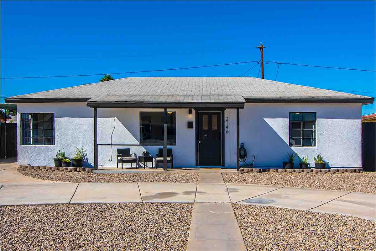 The front view of a single-story home showcases a welcoming entrance with a covered porch. Two chairs are positioned beside the door, flanked by potted plants. A spacious gravel area is visible in front, providing ample parking space.