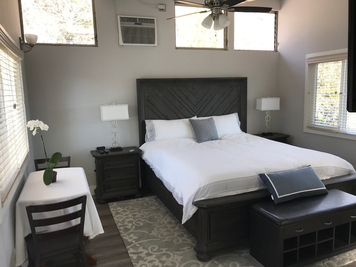 A spacious sleeping area contains a king size bed with crisp white linens and a decorative throw pillow. Two windows allow natural light to fill the room. A small table and chair pair is positioned near the window, with a decorative plant accentuating the space.