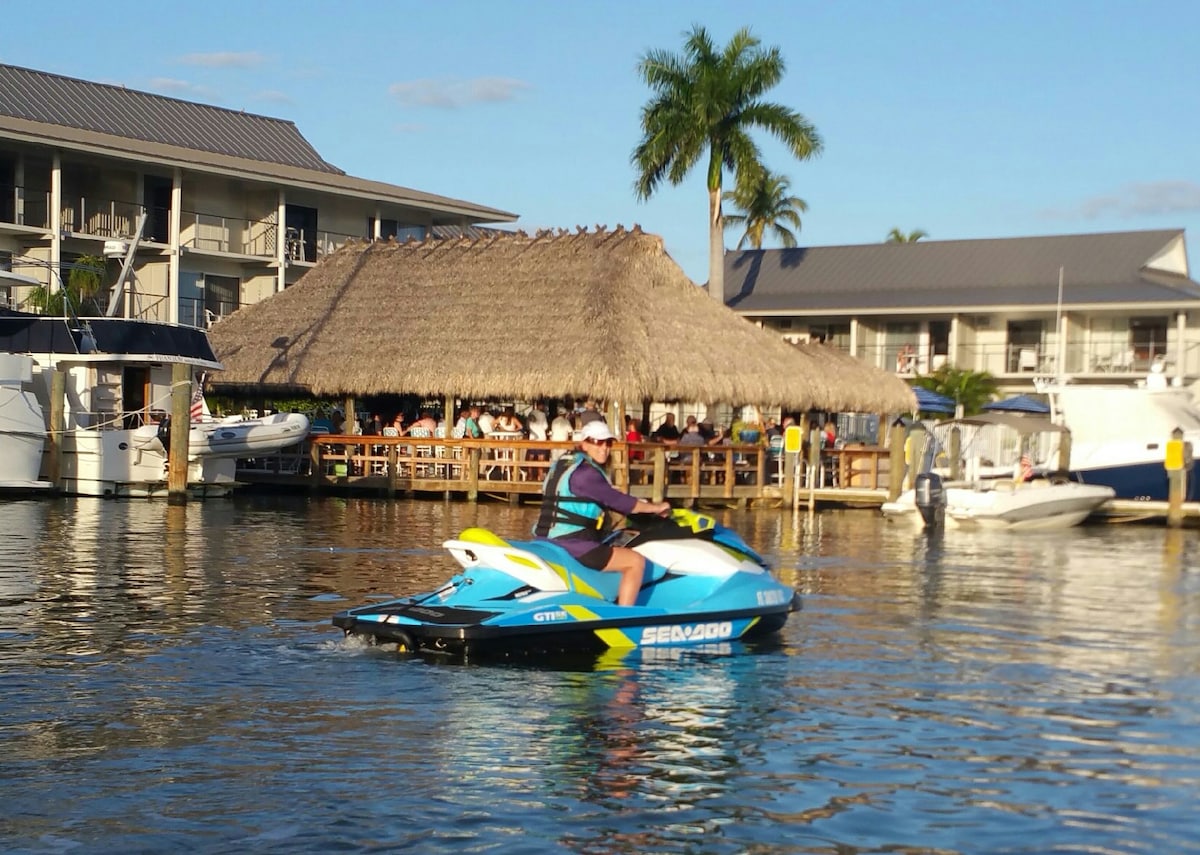 A jet skier is seen navigating the water near a thatched-roof hut, which houses an outdoor dining area. Boats are docked nearby, and palm trees provide a tropical ambiance against a clear blue sky.