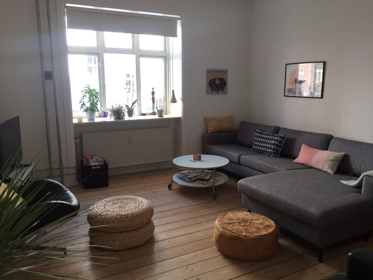 A bright living area features large windows allowing natural light to fill the space. A comfortable grey sectional sofa is complemented by pastel cushions and a round coffee table at the center. Two woven poufs and a potted plant enhance the welcoming atmosphere.