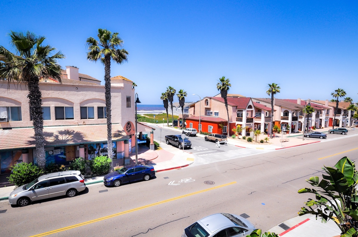 A view of Seacoast Drive showcases palm trees lining the street, with the ocean visible in the distance. Colorful buildings house various shops and eateries, while vehicles are parked along the side of the road under clear blue skies.