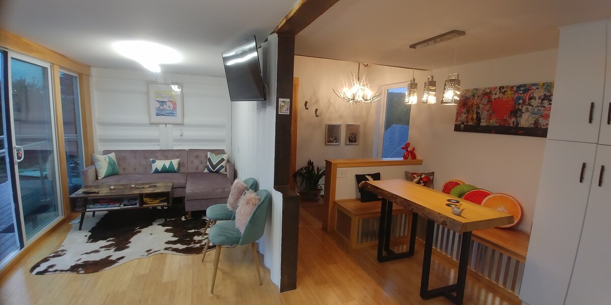 A modern interior is divided into a living area and dining space. A plush couch features colorful cushions, and a dining table is set with vibrant plates. Natural light filters in through large glass doors, illuminating the space and highlighting artistic decor on the walls.