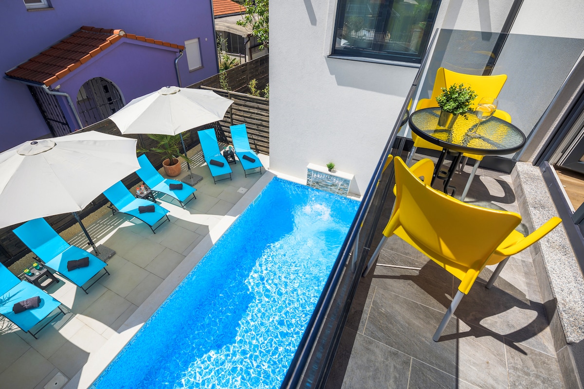 A shared pool surrounded by light blue sun loungers and white umbrellas is visible from the balcony. A small round table and bright yellow chair are situated on the balcony, providing a view of the pool area and nearby greenery.