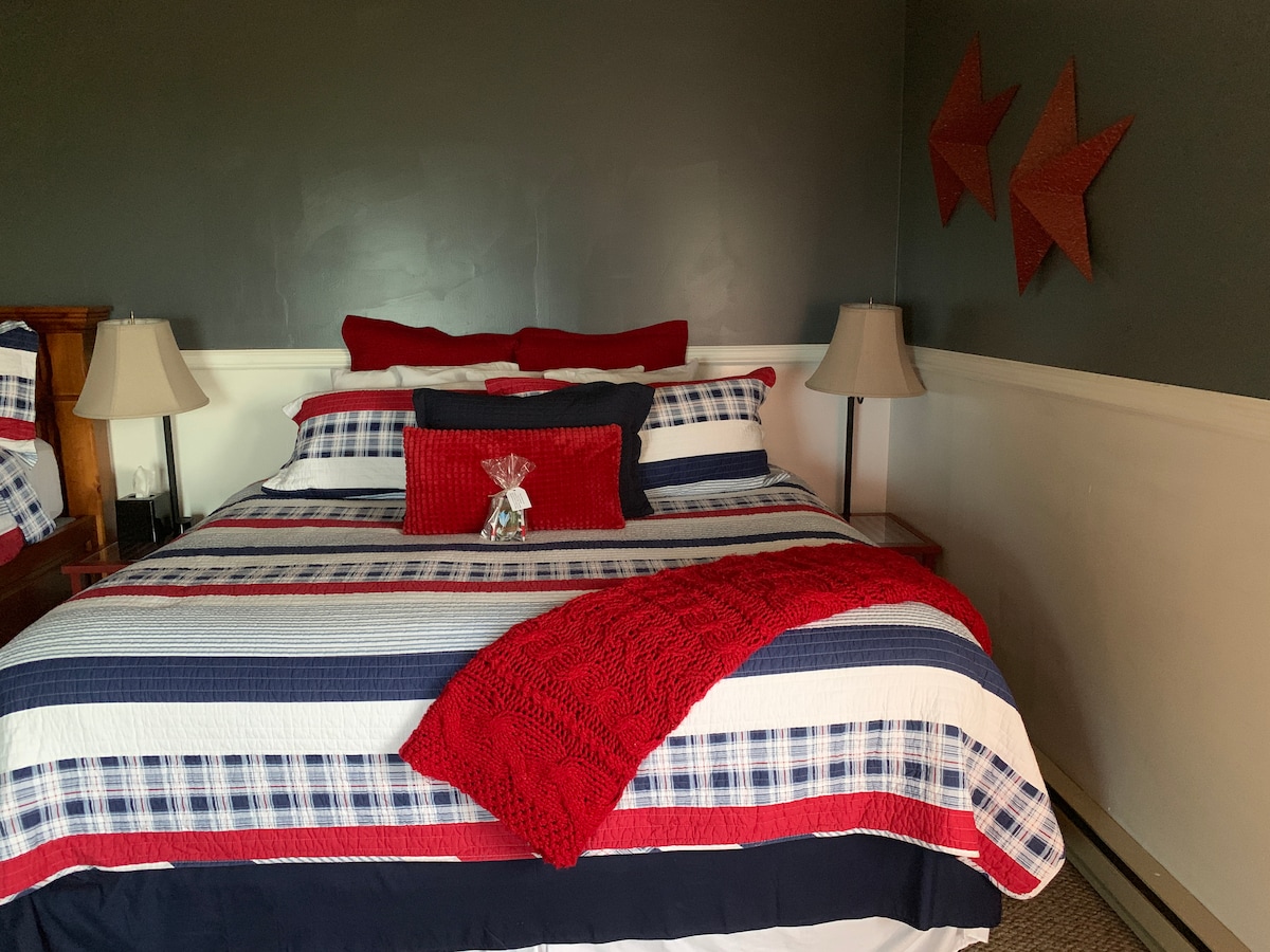 A cozy bedroom features a spacious bed adorned with a striped blanket and red accents. Nightstands with lamps are placed on either side of the bed. The walls are painted a soft gray, complemented by decorative star wall art.