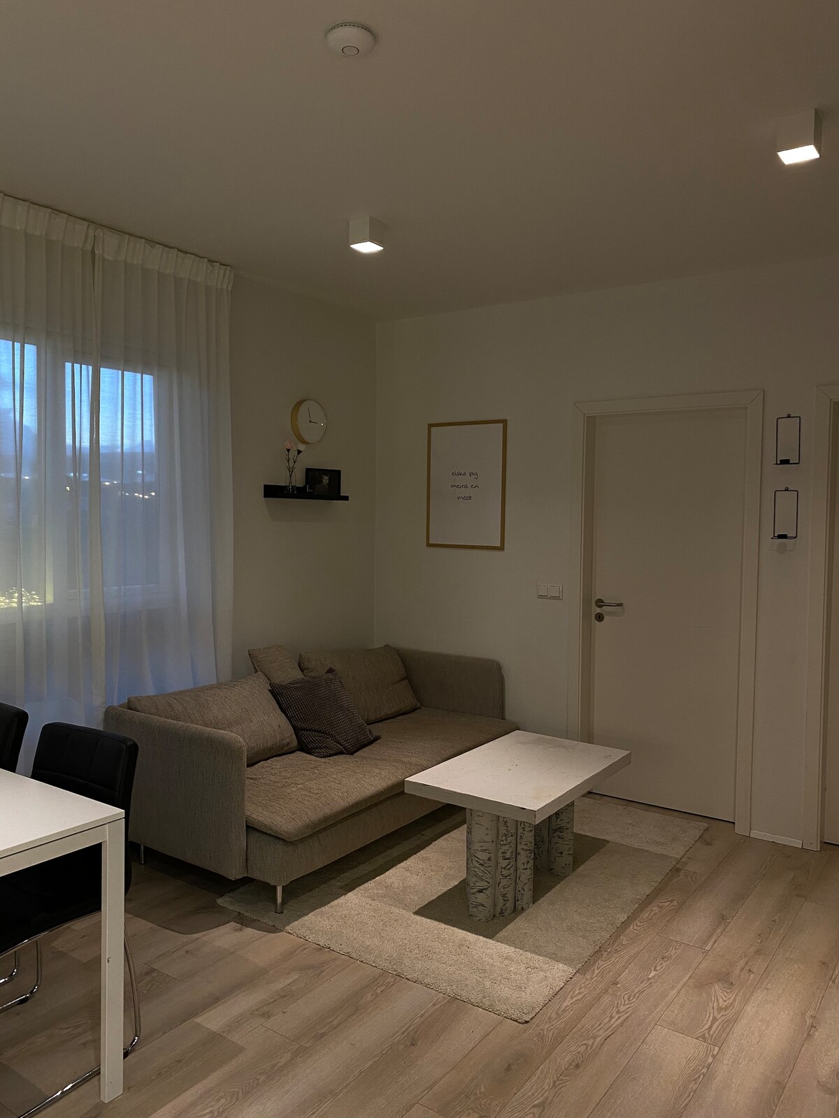 A living area features a light grey sofa with decorative pillows, positioned next to a rectangular coffee table. A small shelf with a clock and decor is mounted on the wall. Natural light enters through a window, complemented by sheer curtains.