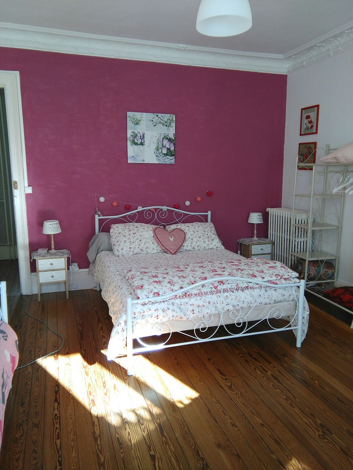 A cozy bedroom features a white metal bed adorned with floral bedding and a heart-shaped decorative pillow. Pink walls add a warm touch, complemented by bedside tables with lamps. A window casts natural light across the polished wooden floor, creating a bright and inviting atmosphere.