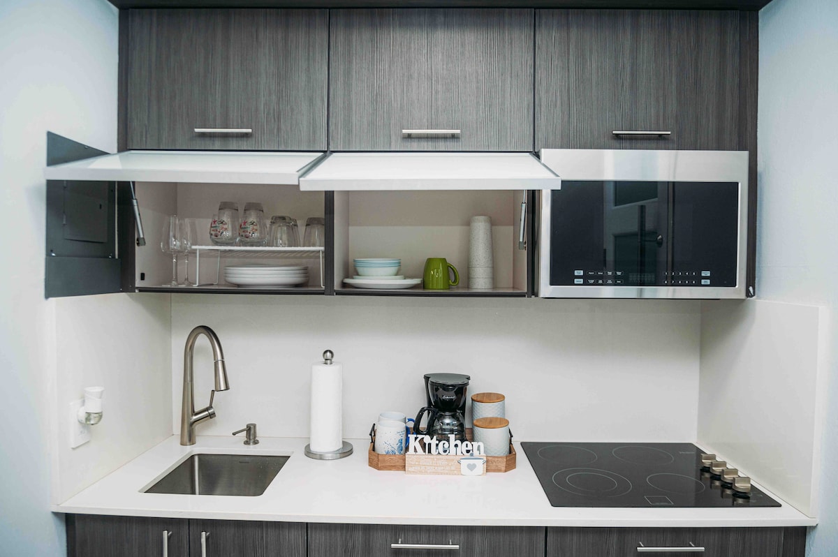 A compact kitchen is equipped with modern appliances including a coffee maker and microwave. Upper cabinets hold glassware and dishware, while a countertop provides a sink and a stove with four cooking burners. Neutral tones and sleek cabinetry create an efficient cooking space.