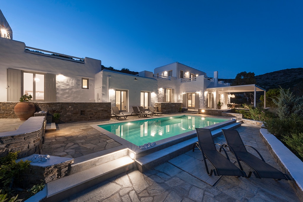 The exterior of the villa is captured at dusk, highlighting a serene outdoor swimming pool surrounded by lounge chairs. Soft lighting illuminates the stone terrace and pathways, creating a welcoming ambiance. The architecture presents a blend of modern and traditional design elements, enhancing the overall privacy.