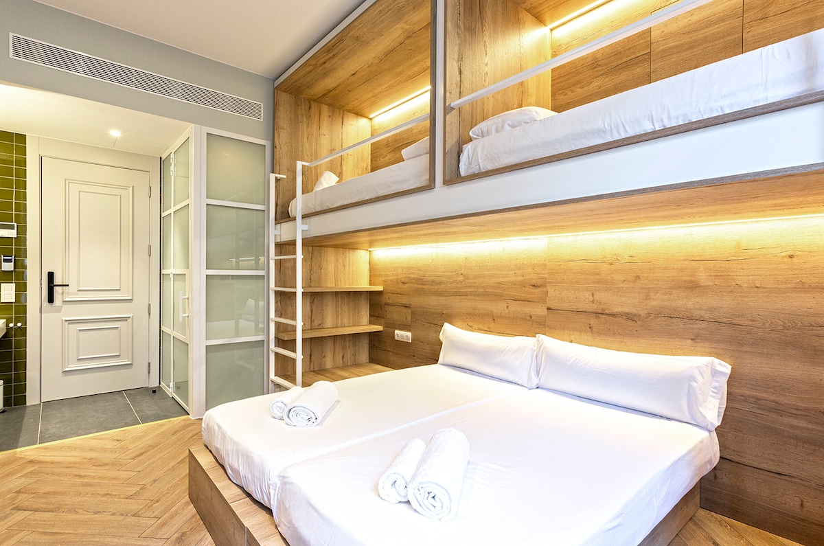A spacious room featuring twin beds that can be arranged as a double or remain as twins. Above, two additional single beds are positioned in a modern design. Soft lighting highlights the wooden walls, while fresh linens and towels are neatly placed on the lower beds.