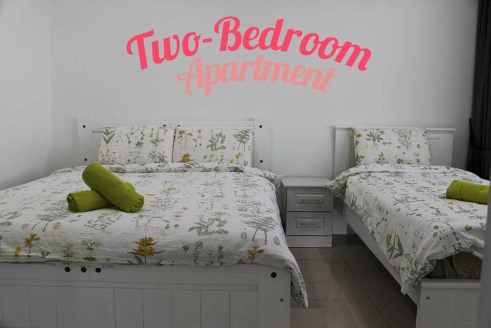 The room features two beds, one queen and one single, both adorned with floral-patterned bedding. Green towels are neatly arranged on each bed. The walls are painted white, and a decorative sign reads 'Two-Bedroom Apartment' in vibrant pink.