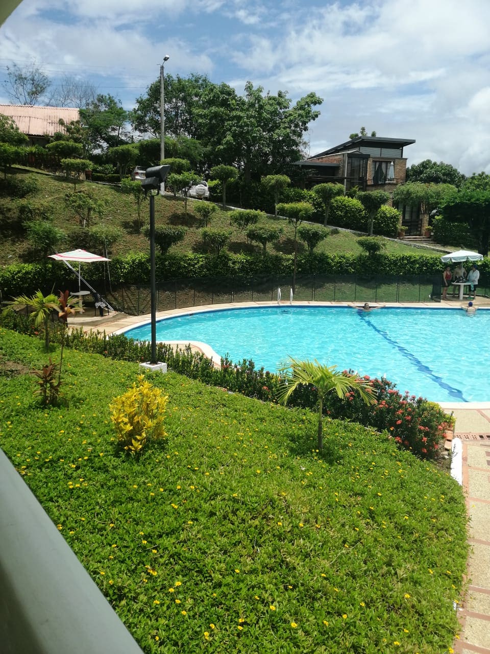 A clear swimming pool is surrounded by well-maintained greenery, featuring manicured grass and flowering plants. A shaded seating area is visible beside the pool, with gentle slopes leading to the backdrop of trees and additional structures in a peaceful setting.