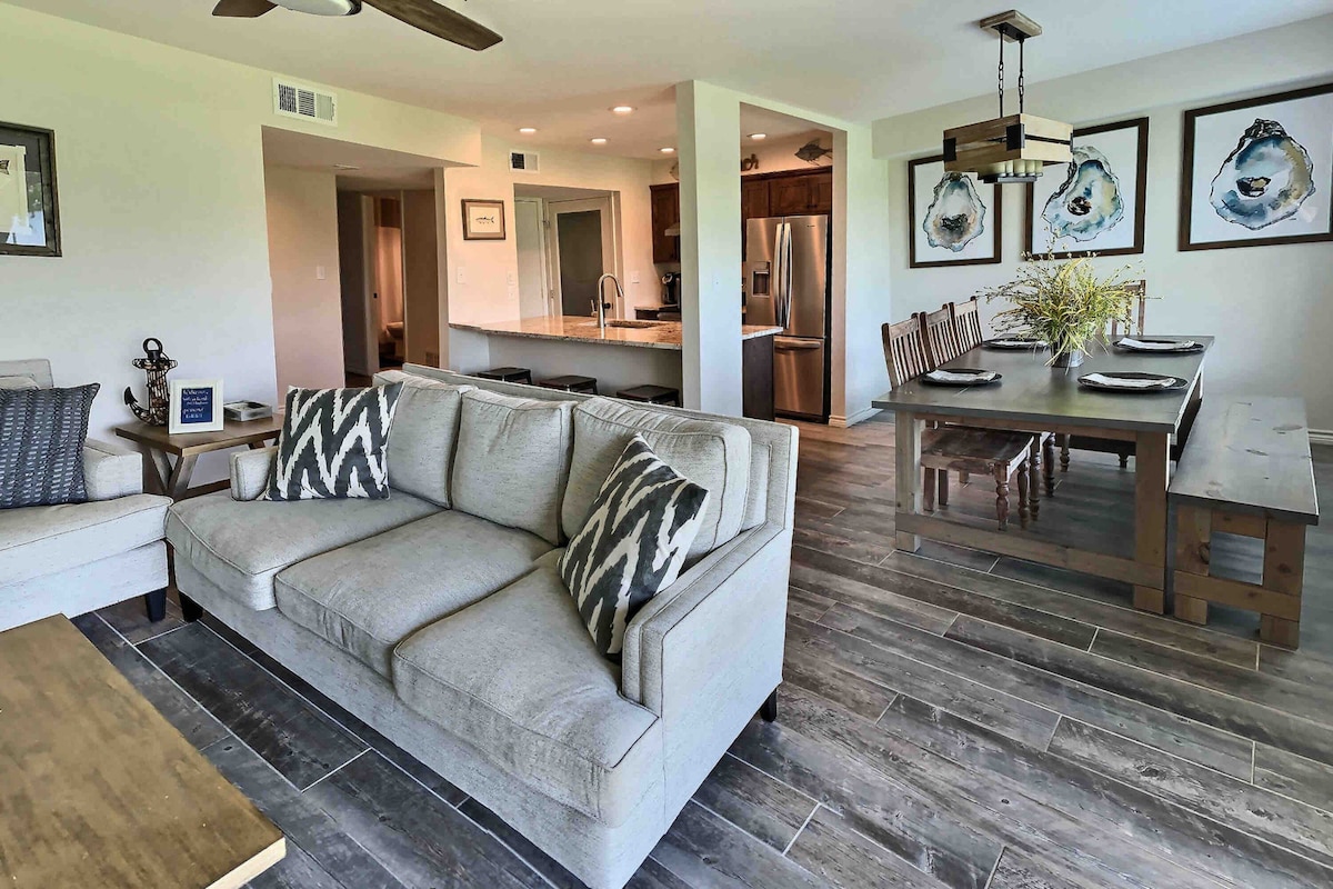 An open-concept living area features a light gray sectional sofa with patterned cushions, complemented by a wooden coffee table. A dining area includes a long wooden table surrounded by chairs, with decorative wall art displayed nearby. Natural light flows in, enhancing the spacious feel.