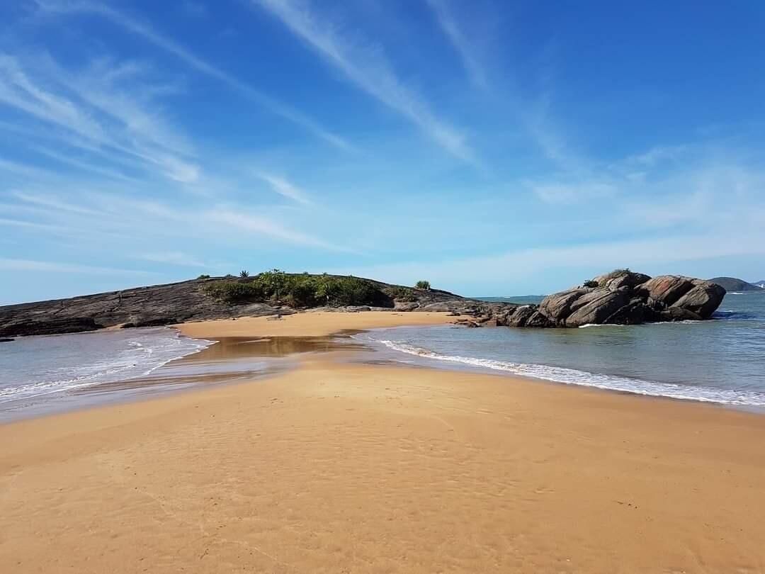 A serene beach scene features soft golden sand meeting gentle waves. Lush greenery is visible in the distance, while rugged rocks outline the shoreline. The expansive blue sky is decorated with wispy clouds, creating a sense of openness and tranquility.