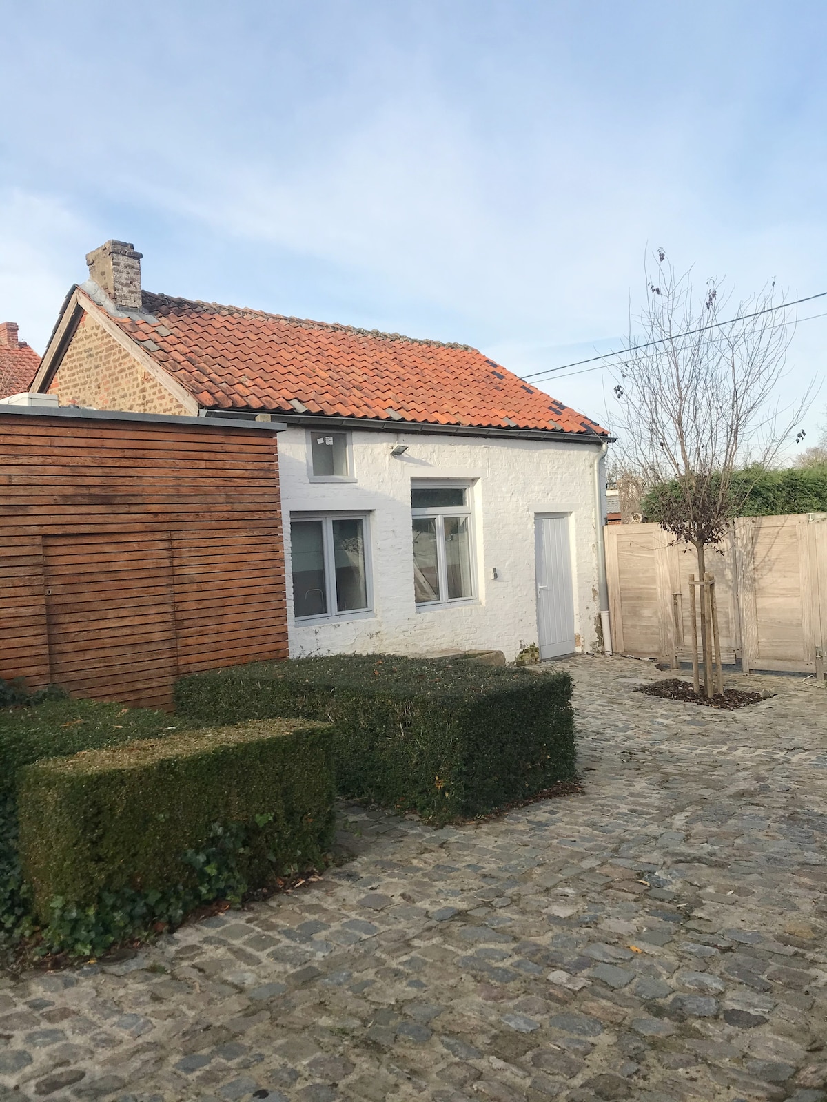 The exterior of the cottage features a charming blend of white walls and a red-tiled roof. A wooden fence and neatly trimmed hedges guide the entrance, while a stone pathway leads to the door, contributing to the serene ambiance of the surrounding area.