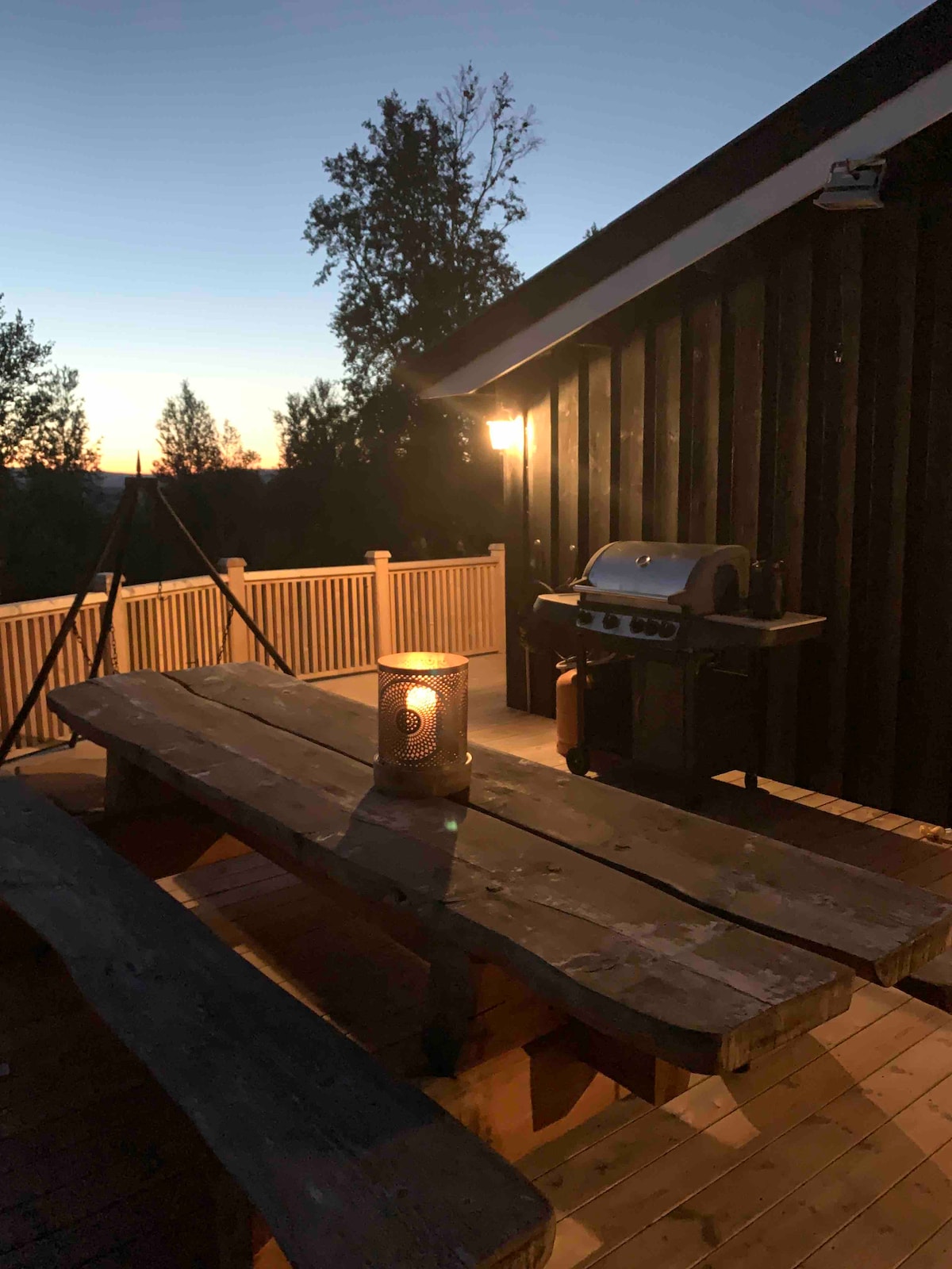 An outdoor deck features a rustic wooden picnic table, illuminated by a candle lantern. A barbecue grill is positioned nearby, and the surrounding trees silhouette against a twilight sky, enhancing the serene evening ambiance.
