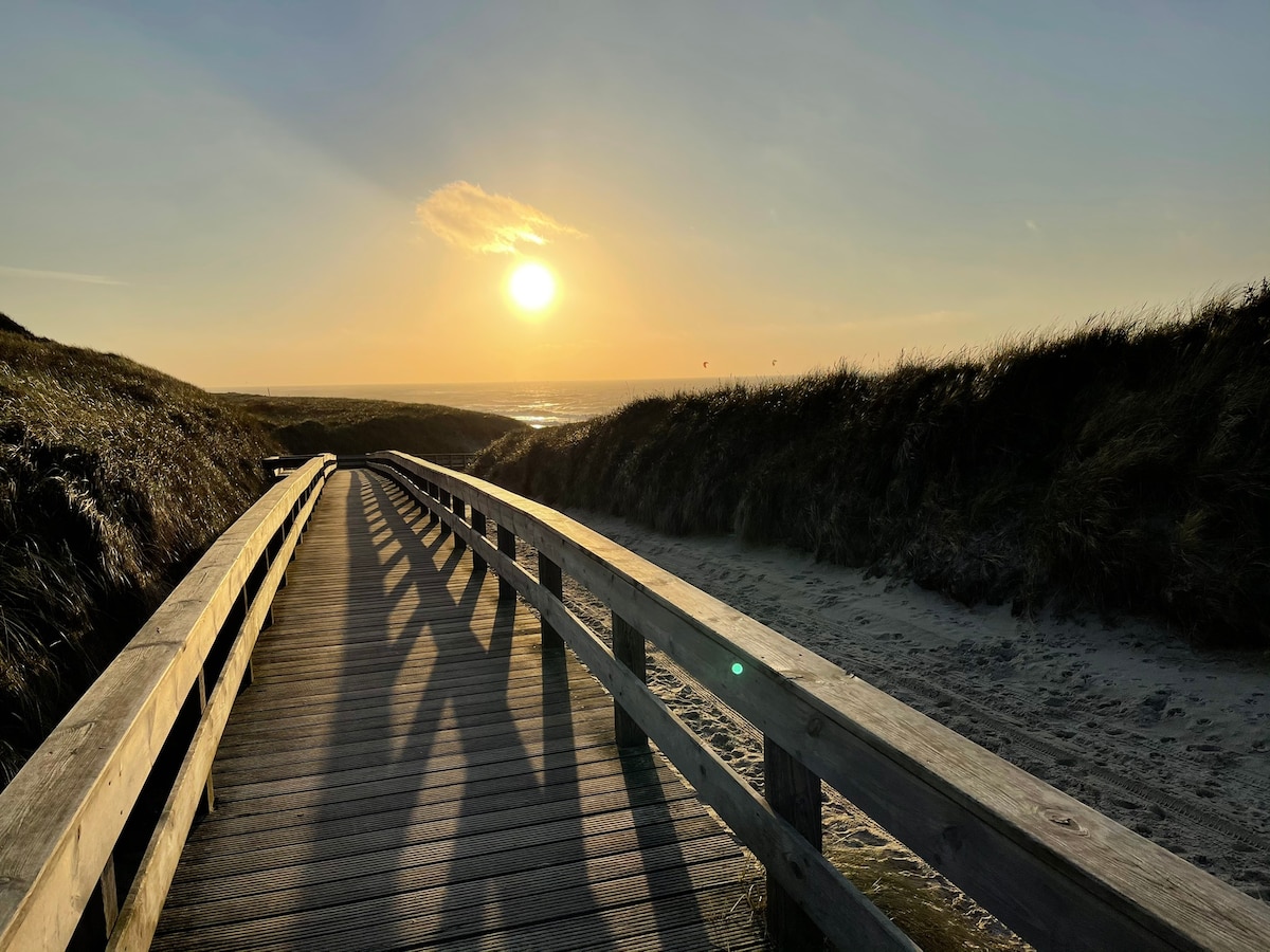 A wooden boardwalk meanders through grassy dunes, leading toward a serene beach at sunset. The sun casts a warm golden light over the scene, reflecting on the gentle waves of the ocean in the distance.