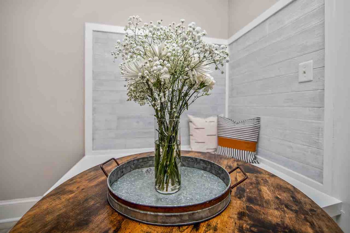 A glass vase filled with white flowers is positioned on a round, weathered wood table. The table is set on a rustic tray, enhancing the natural aesthetic. Neutral-colored cushions are visible in the background, adding subtle texture to the room.