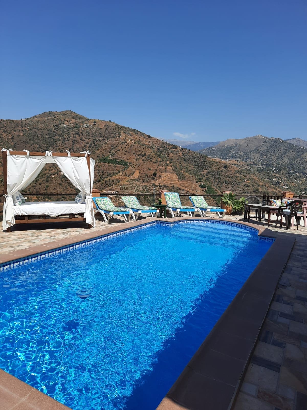 A private swimming pool glistens under the clear blue sky, surrounded by mountains. Sun loungers provide a relaxing space for sunbathing, while a shaded area with draped fabric offers a comfortable retreat. The scene captures a serene outdoor environment ideal for leisure.
