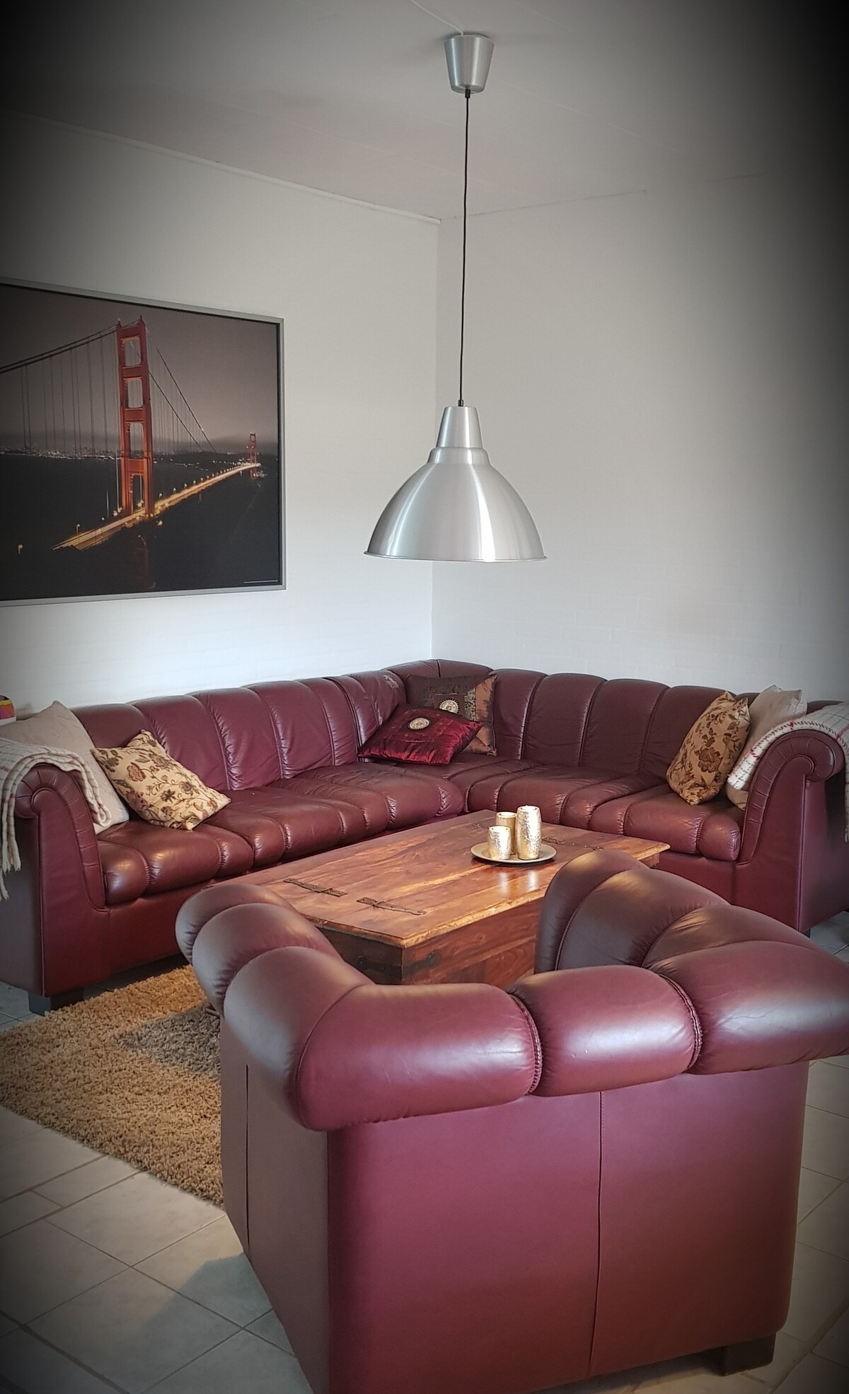 A spacious living area includes a deep red leather sectional sofa surrounding a wooden coffee table. Decorative pillows add a touch of comfort. A large framed photograph of the Golden Gate Bridge hangs on the wall, and a stylish pendant light illuminates the space.