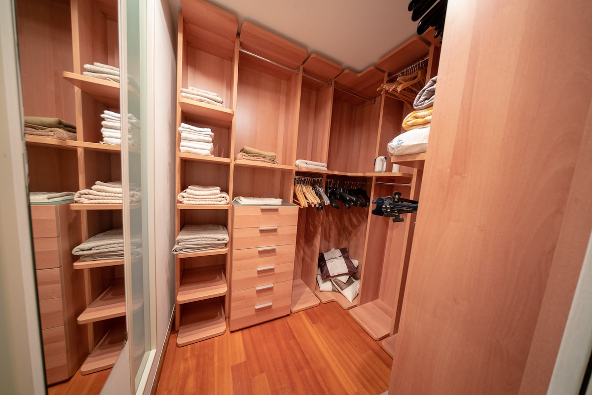 A well-organized walk-in closet features wooden shelving and drawers. Shelves are stocked with neatly folded towels and linens, while clothing hangers are available for guest apparel. A mirror reflects the space, enhancing the sense of openness and utility.