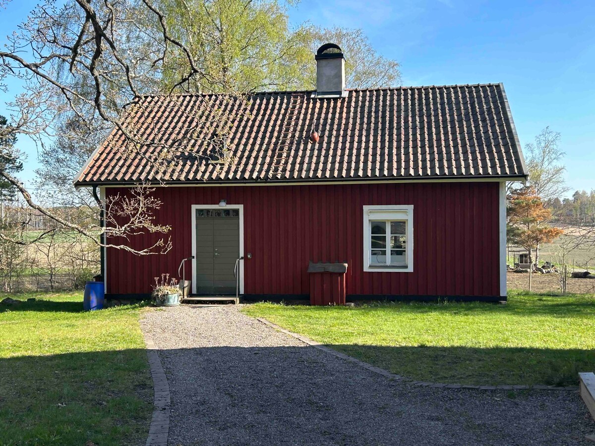 A red wooden cottage is seen from the front, with a traditional pitched roof and a chimney. A gravel path leads to the entrance, bordered by a grassy area. Nearby, a tree provides shade, and the landscape features distant fields and greenery.