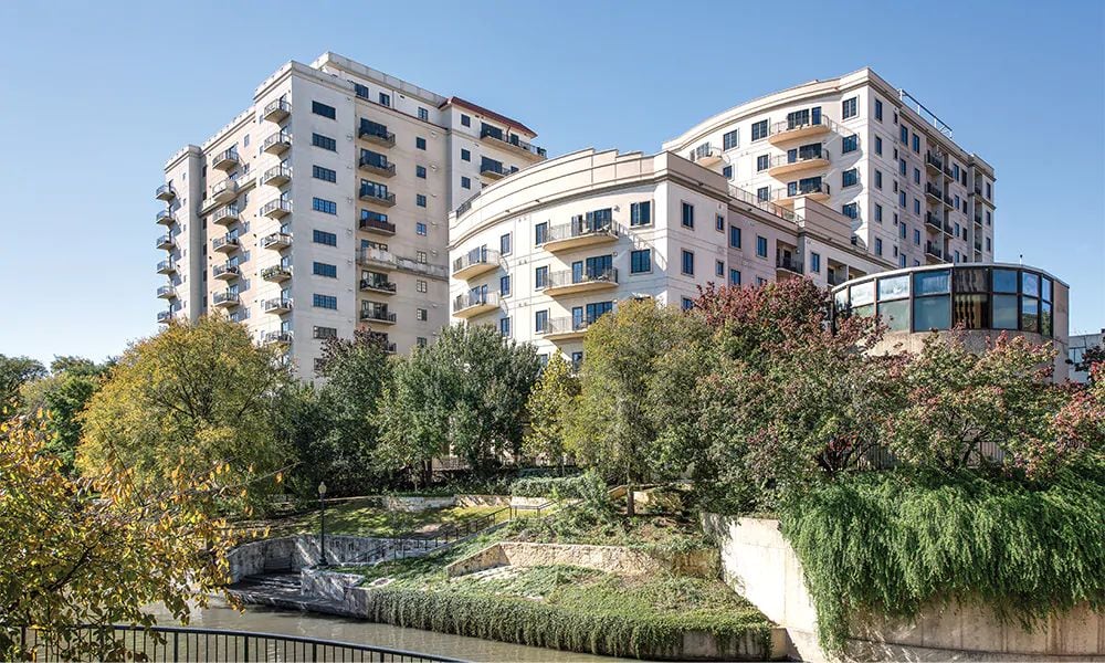 The resort building is prominently featured alongside lush greenery and a flowing waterway, showcasing modern architecture with balconies. The arrangement of trees and shrubs adds texture to the landscape, creating a harmonious blend between the building and its natural surroundings.