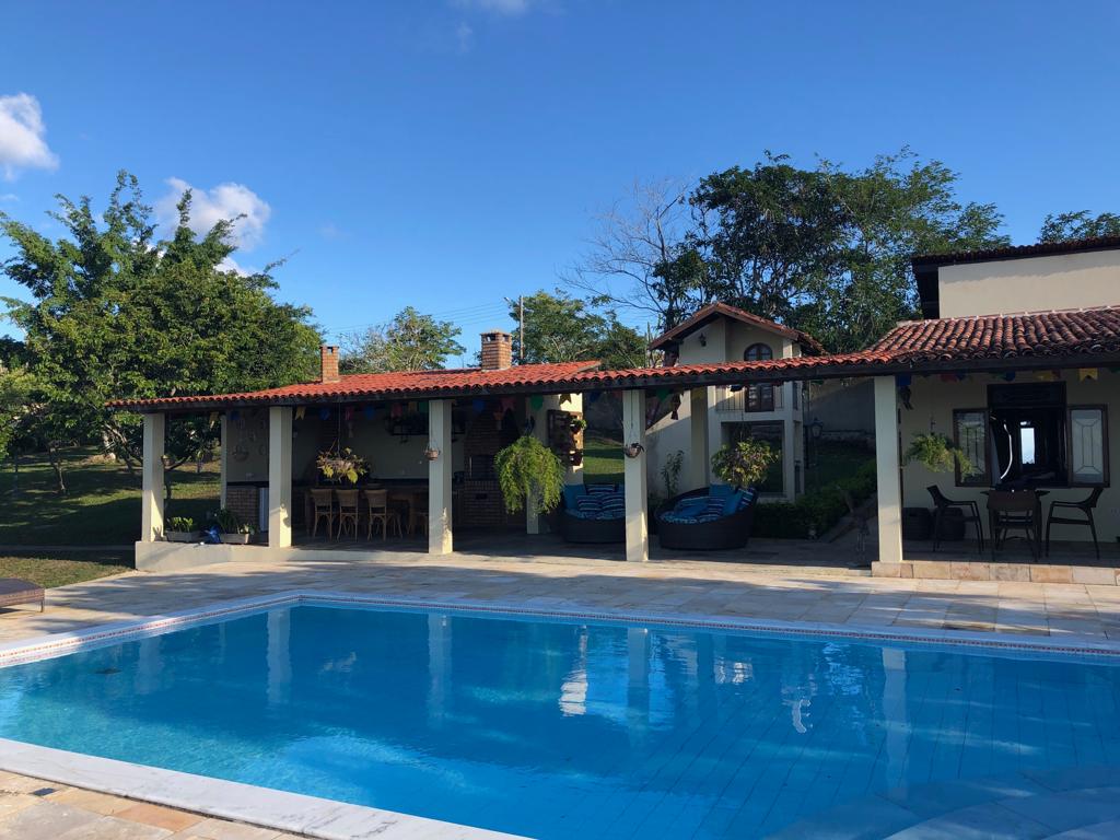 A spacious outdoor area showcases a bright blue pool surrounded by a stone patio. Covered seating areas with wooden tables and chairs are positioned near the house, which features a rustic design. Lush greenery provides a natural backdrop, enhancing the sense of tranquility.
