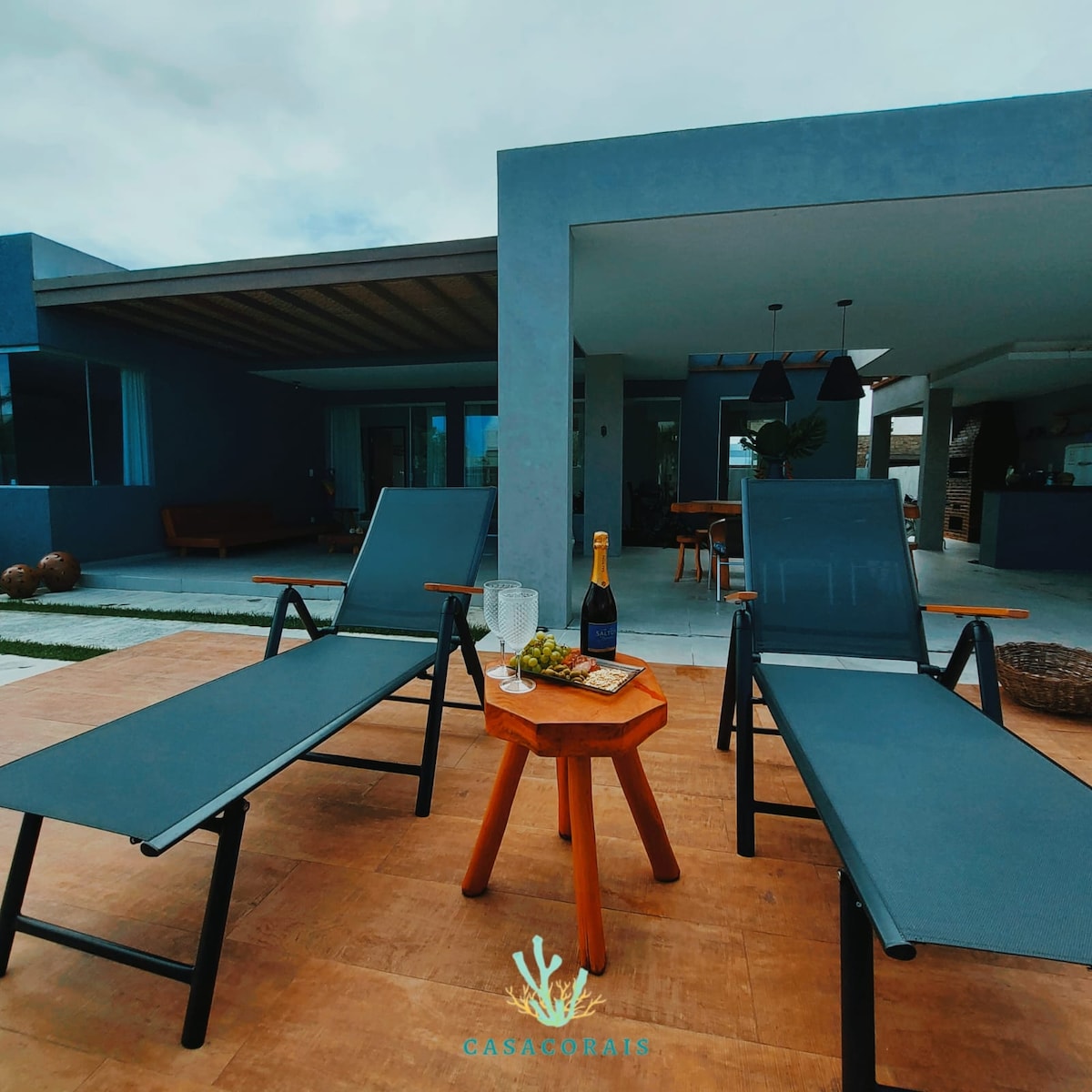 Two lounge chairs are arranged on a wooden deck, positioned thoughtfully next to a small wooden table. The table holds a bottle of champagne and two glasses. In the background, a modern, blue house with large windows and an inviting outdoor area is visible.