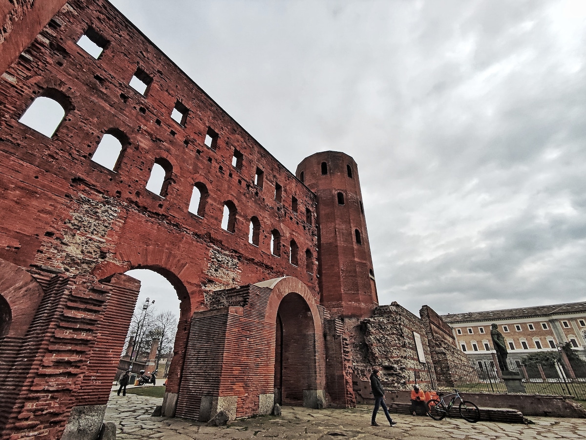 An ancient brick structure features large arched openings and a round tower, showcasing remnants of its historical significance. The textured walls exhibit variations in clay and stone. Overcast skies provide a muted backdrop to visitors exploring the area around the ruins.