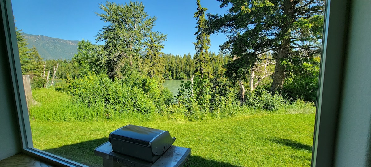 A clear view of a verdant landscape showcases tall trees and lush grass. Sunlight enhances the vibrant greenery, with a glimpse of a distant body of water visible through the trees. A patio area is indicated by a covered surface in the foreground.