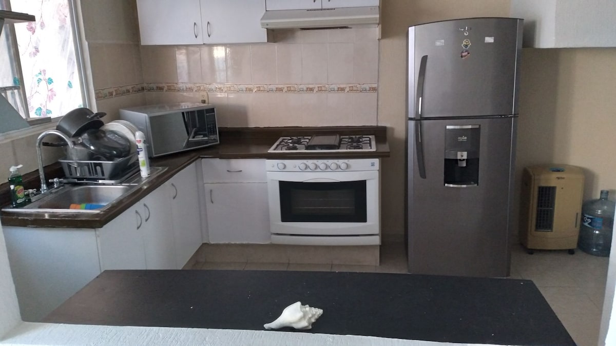 A functional kitchen area is presented with essential appliances, including a stove, oven, microwave, and refrigerator. Counter space is utilized effectively, with a sink and dish drying rack visible. Cabinets are arranged above and below the counter, enhancing storage options.