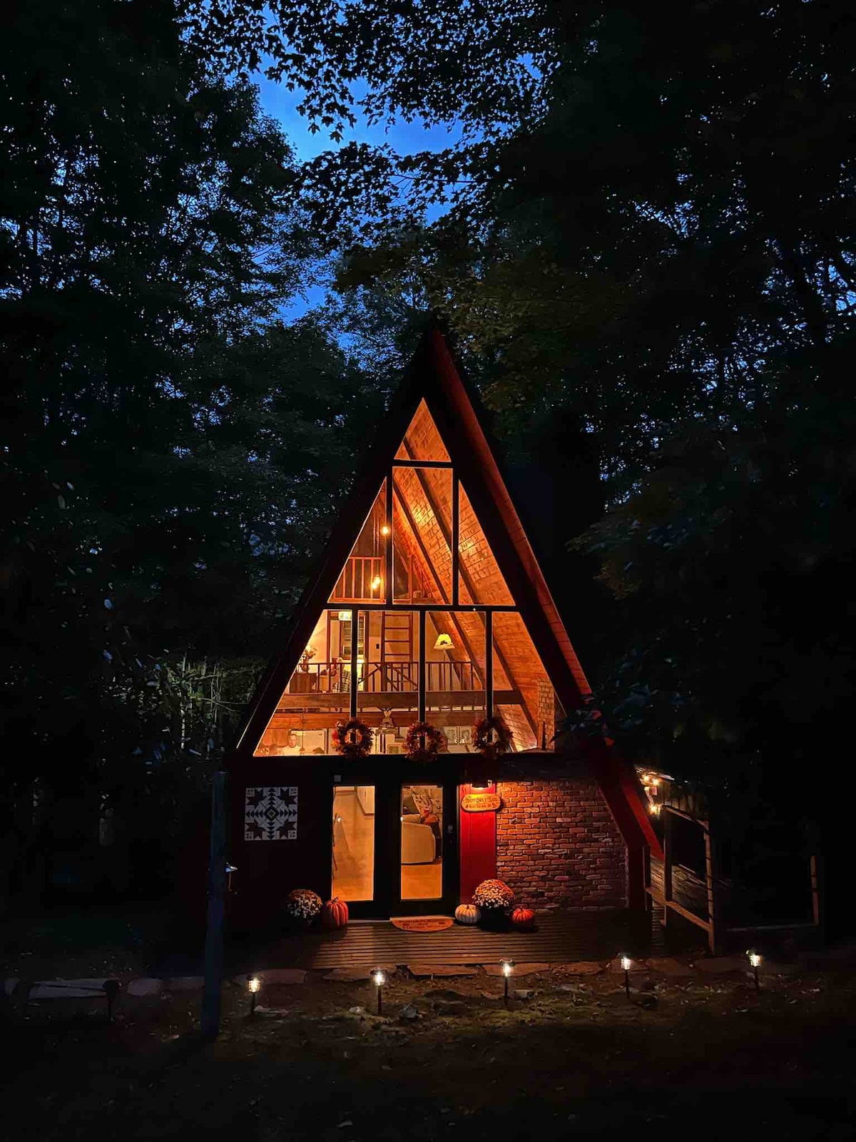 An A-frame cabin is showcased at dusk, surrounded by trees. Warm light glows from large windows, revealing a cozy interior. The entrance features a welcoming porch with decorative elements. Soft lighting along the pathway creates an inviting atmosphere in the natural setting.