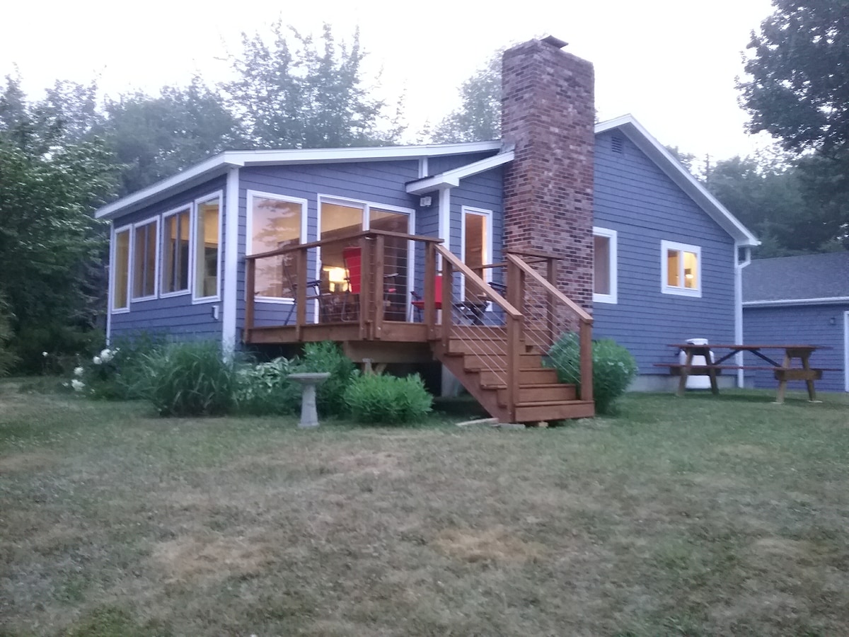 The cottage features a blue exterior with a prominent stone chimney. A spacious wooden deck with steps leads to the entrance. Large windows provide views of the surrounding greenery. A picnic table is positioned nearby on the lawn, creating a welcoming outdoor space.