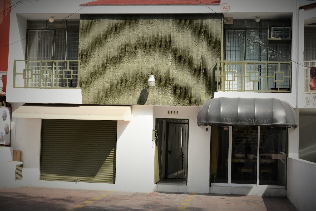 The exterior of a two-story building features a textured green facade, with a covered entryway to the side. A wide, retractable awning extends over the entrance, and large windows provide natural light. A small balcony is visible on the upper level.