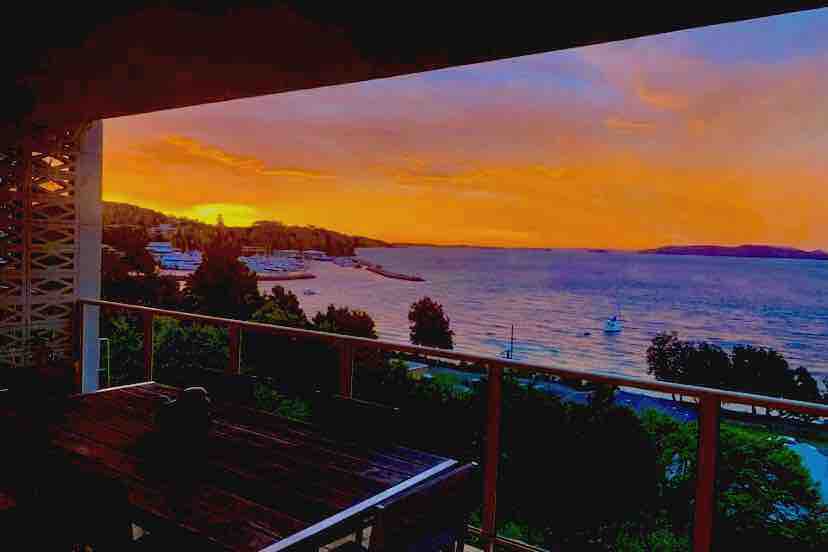 An elevated balcony offers an expansive view of the water at sunset, with vibrant hues of orange and purple in the sky. Nearby, the coastline is visible, accented by boats and lush greenery. A dining table is positioned on the balcony, enhancing the outdoor experience.