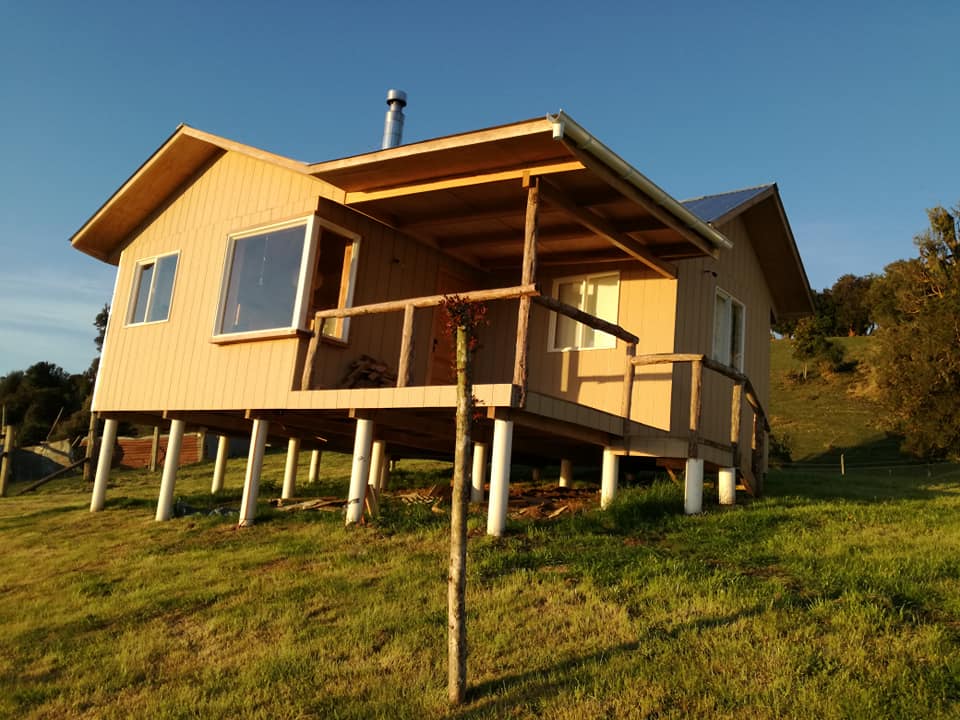 A charming wooden cabin is positioned on stilts, featuring large windows that provide expansive views of the surrounding landscape. The structure is set against a grassy area, with a gentle slope leading to the hillside. A chimney is visible on the roof, indicating a cozy indoor atmosphere.
