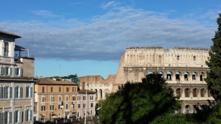 A view of the Colosseum can be seen alongside charming buildings featuring classic Roman architecture. The sunny sky highlights the historic stone structure, while lush greenery adds a touch of nature to the urban landscape.