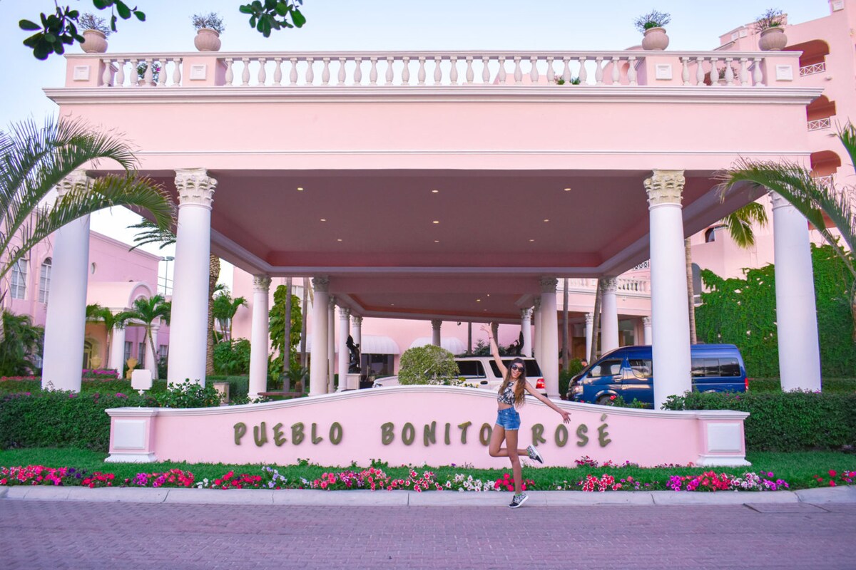 The entrance to a resort is framed by light pink walls and adorned with vibrant flower beds. An elegant portico is supported by white columns, offering a welcoming ambiance. The sign displays 'Pueblo Bonito Rose' prominently, inviting guests into the property.