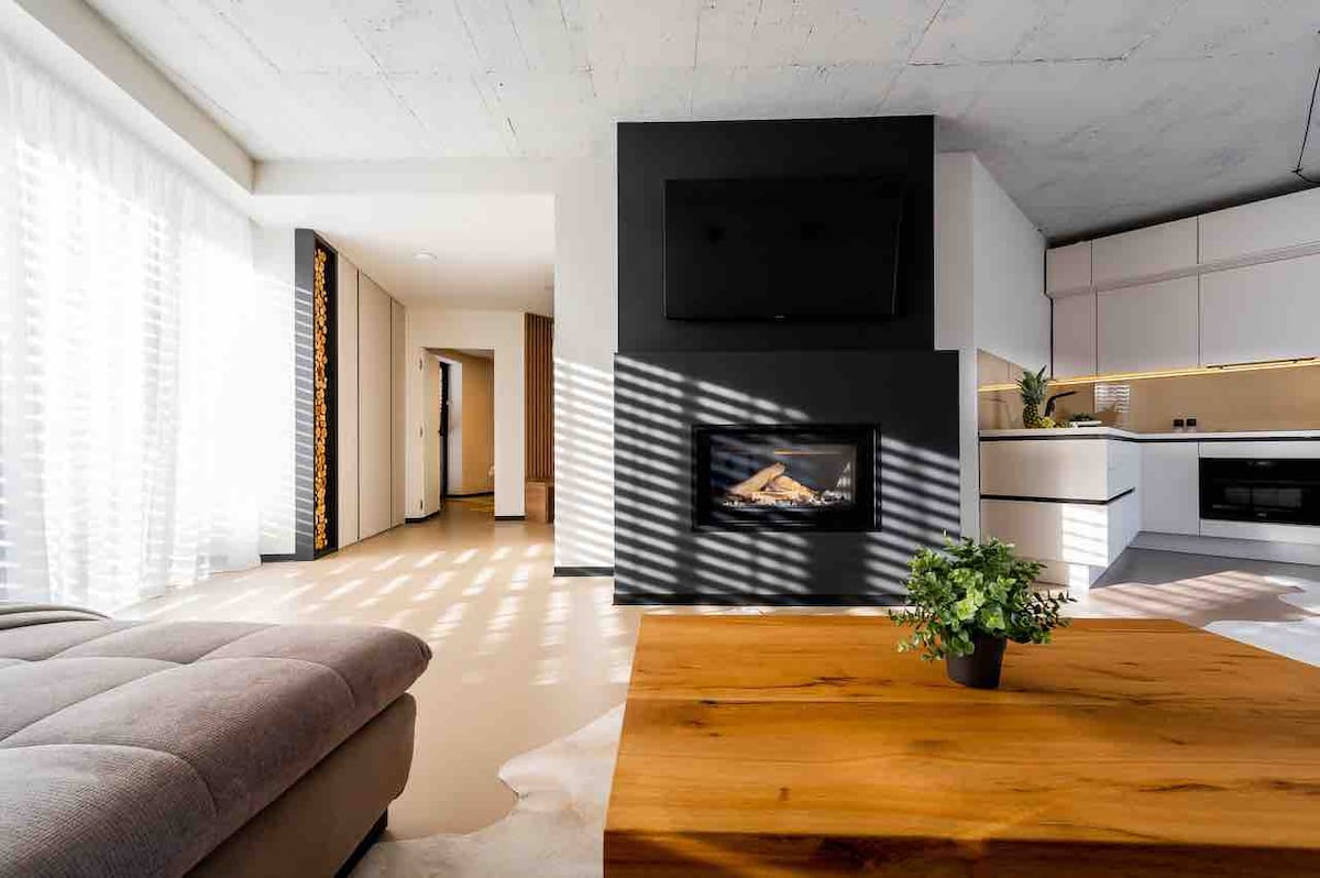 A modern living area features a spacious grey sectional sofa and a wooden coffee table with a small potted plant. Light streams in through sheer curtains, creating a warm ambiance. A sleek fireplace is mounted on a black accent wall, with a flat-screen television above.