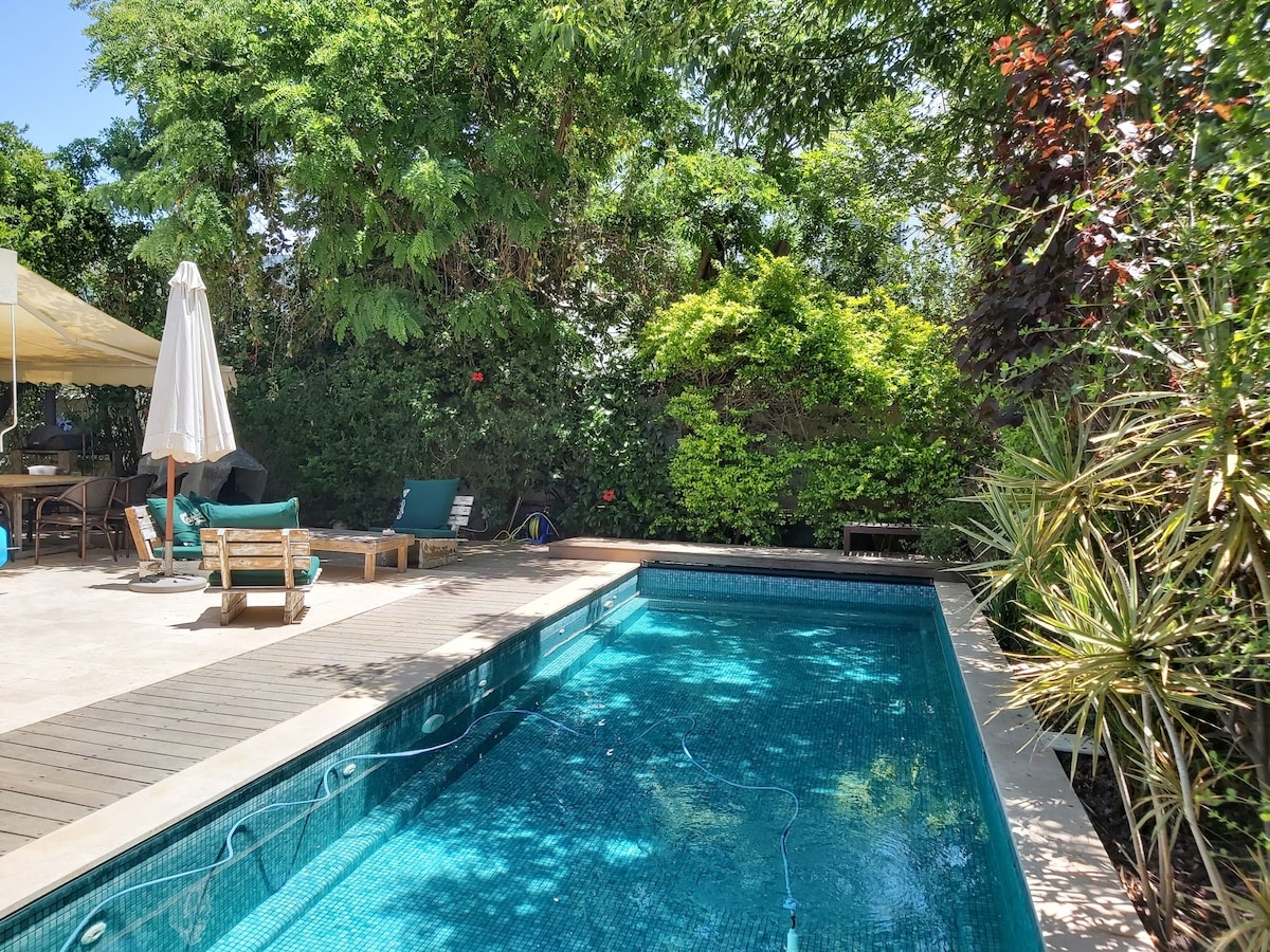 A large, private pool is surrounded by lush greenery, providing a natural backdrop. Sun loungers are positioned on the pool deck, complemented by a shaded seating area under a canopy. The sparkling water invites relaxation in this serene outdoor space.