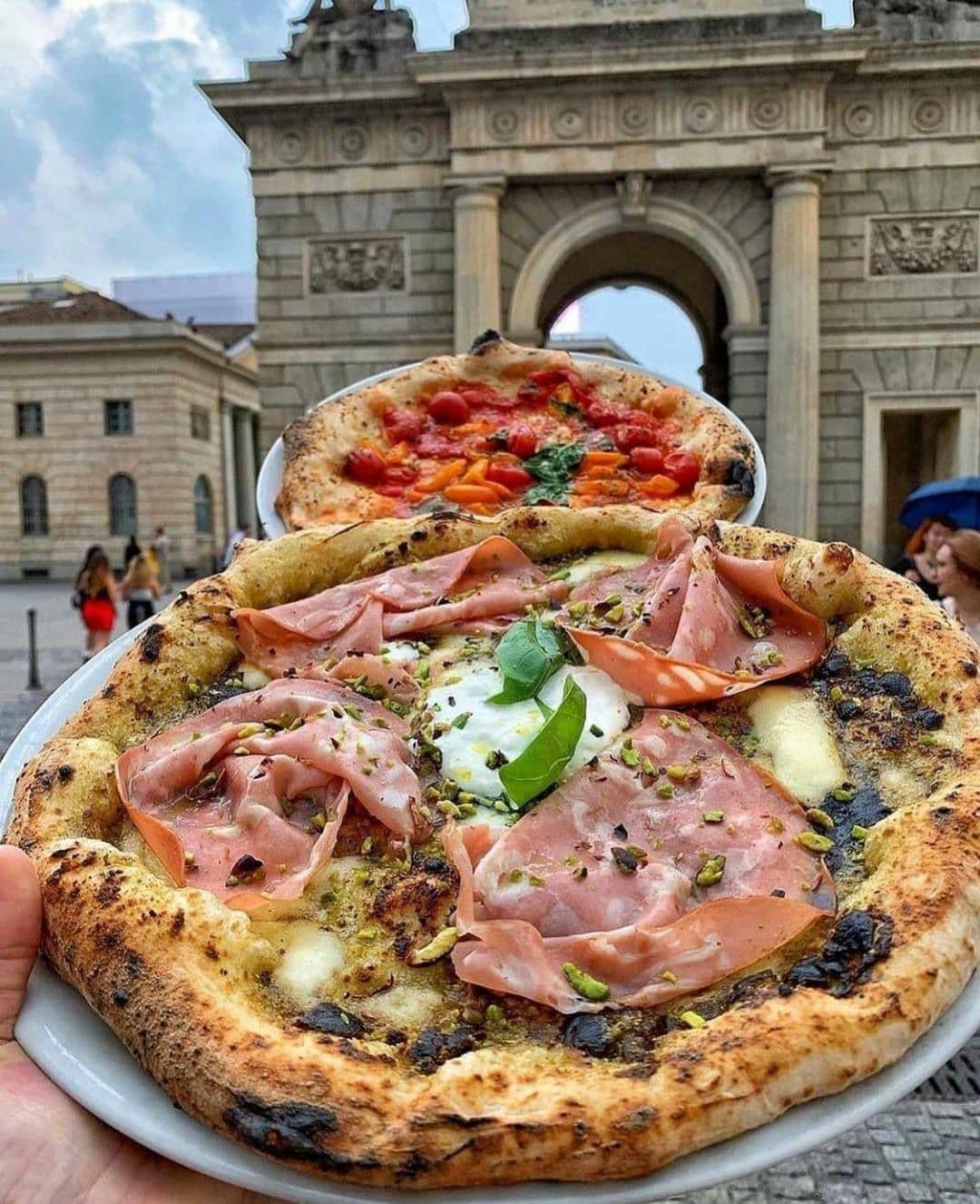 Two pizzas are presented, showcasing their rustic, charred crusts. The pizza on the bottom features melted cheese, slices of ham, and garnished with green basil. The top pizza is topped with fresh tomatoes. The backdrop includes an architectural archway, adding context to the dish.