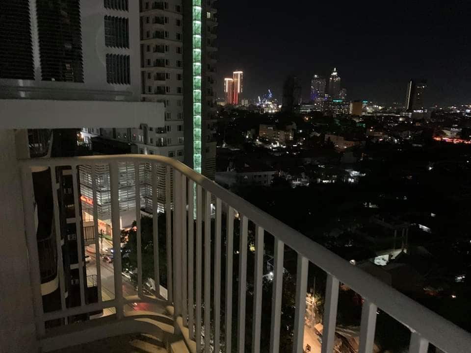 A balcony offers a view of the city skyline at night, showcasing illuminated buildings and distant lights. The railing is white, and the surrounding structures provide a sense of height and perspective over the urban area below.