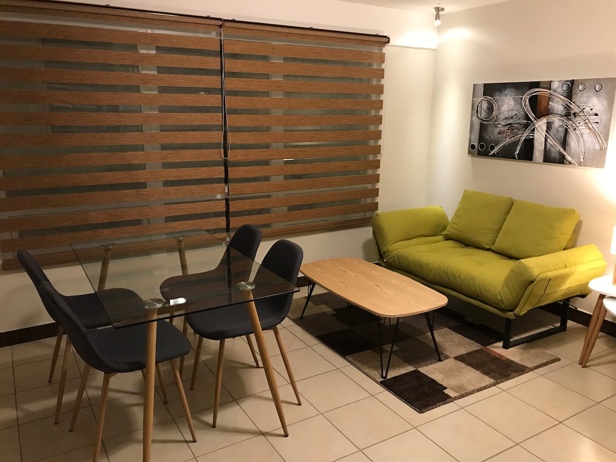 A modern living area is furnished with a bright green sofa and a wooden coffee table. A glass dining table with four chairs complements the space. Dual-textured blinds filter light through a large window, while abstract wall art adds an artistic touch.