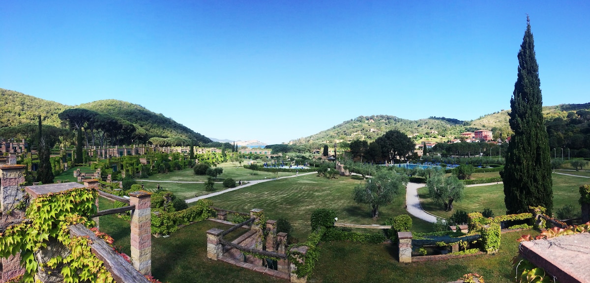 A panoramic view of the surrounding landscape reveals lush green hills, well-maintained lawns, and terraced gardens. Pathways wind through the area, leading to various sections of the gardens. Natural beauty and tranquility are emphasized by the blue sky and distant architectural elements.