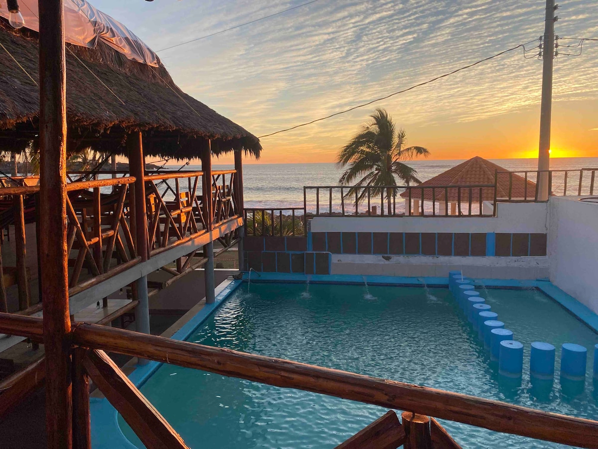 An outdoor area is featured with a swimming pool reflecting the vibrant colors of the sunset. Palm trees gently sway in the breeze, while a thatched-roof structure provides shaded seating overlooking the ocean. The setting creates a serene atmosphere for relaxation.