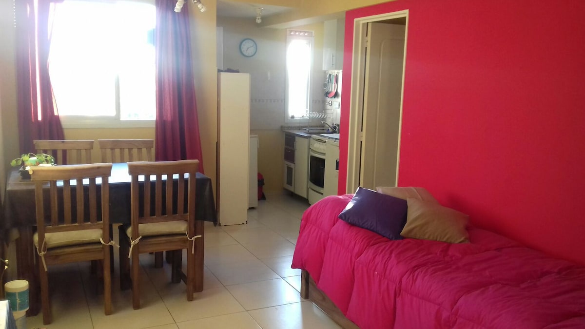 A bright and open living area features a compact dining table surrounded by wooden chairs. A spacious bed with vibrant bedding is positioned nearby. The kitchen area, visible in the background, includes white cabinetry and essential appliances. Natural light filters through the window, enhancing the space.