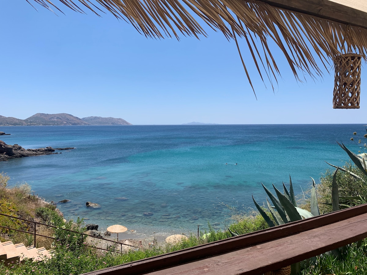 A sweeping view of the serene sea showcases crystal-clear turquoise waters merging with the horizon. Gentle waves lap against rocky shores, while distant hills create a calming backdrop. Green vegetation and natural elements frame the scene, enhancing the coastal ambiance.