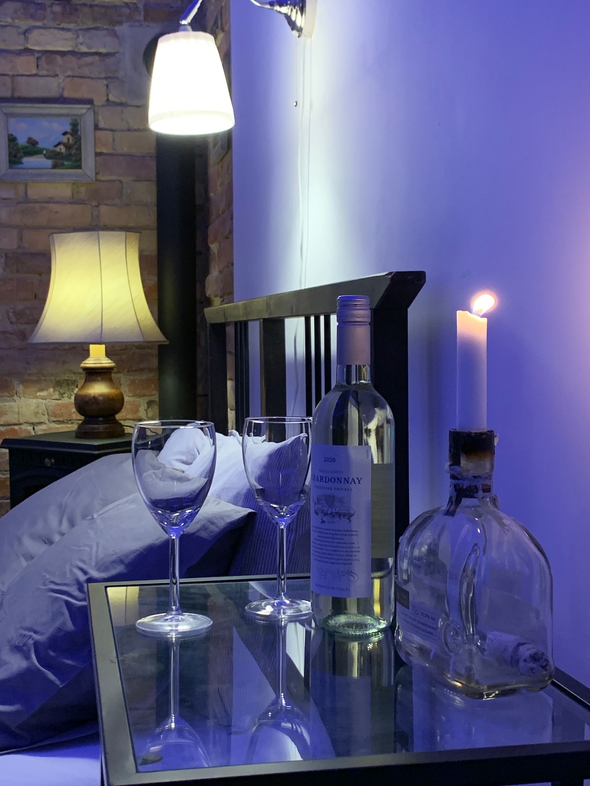 A cozy bedside table is presented with two glasses and a bottle of white wine. A single candle adds soft lighting beside the glass surface. Light fixtures above enhance the space, with a softly glowing lamp and vibrant blue walls complementing the exposed brick backdrop.